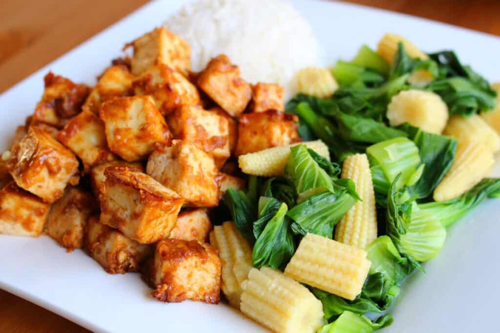 closeup image of tofu coated in peanut sauce with a side of rice and sauteed bok choy with chopped baby corn