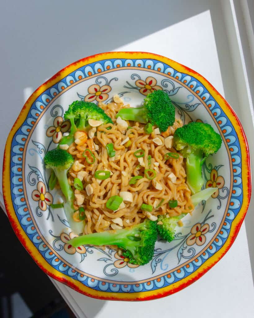 Overhead shot of Peanut Butter Noodles in a bowl. There are florets of broccoli around the outside of the bowl and the noodles are topped with chopped peanuts and scallions.