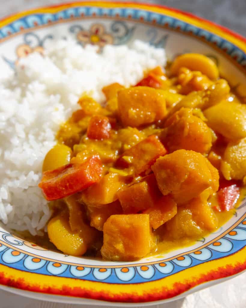 A close up shot of a bowl of Easy Sweet Potato Curry. There are chunks of sweet potato, carrots, red bell pepper, along with butter beans. It is served next to white rice.