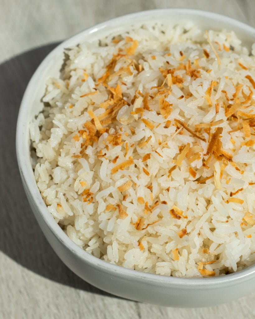 A close up of a white bowl filled to the brim with Coconut Rice. There are golden brown flakes of toasted coconut on top.