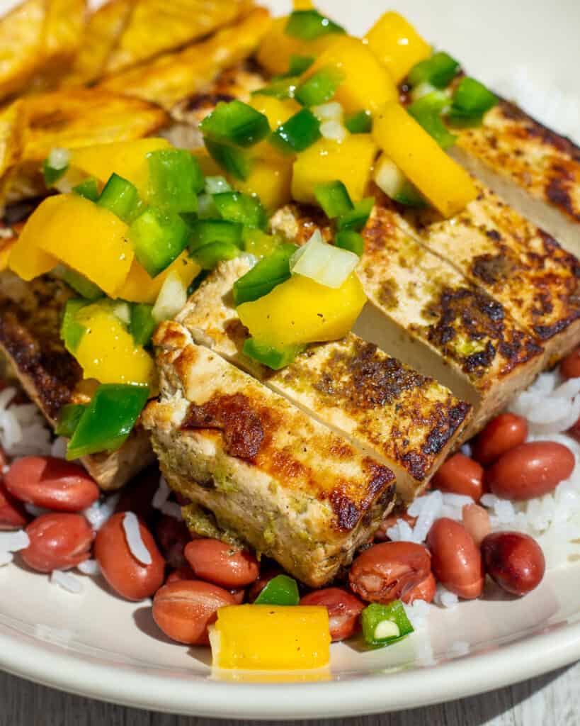 Sliced jerk tofu sitting on a bed of white rice and red beans. It's topped with mango salsa, consisting of mango, jalapeño peppers, and onion.