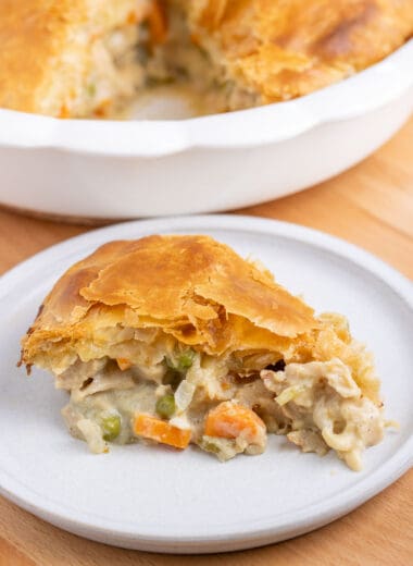 A slice of vegan chicken pot pie on a small white plate. The full baking dish with the pot pie is blurry in the background.